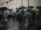 black and white image of passengers standing at a mumbai railway station in the rain