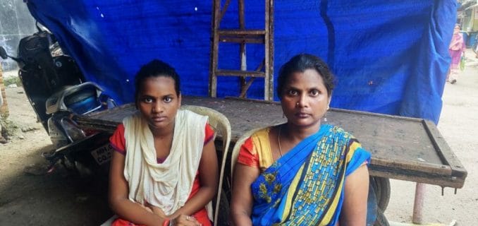 Priya and her mother Laxmi look at the camera.  They sit on two plastic chairs, behind them a blue tarpaulin.